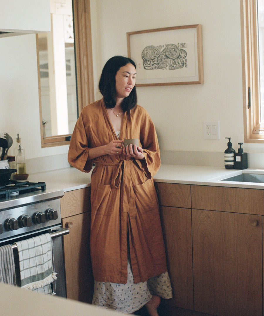 A woman wearing a camel linen robe stands in a kitchen holding a small mug. The kitchen features light wood cabinets, a white countertop, and a framed print on the wall.