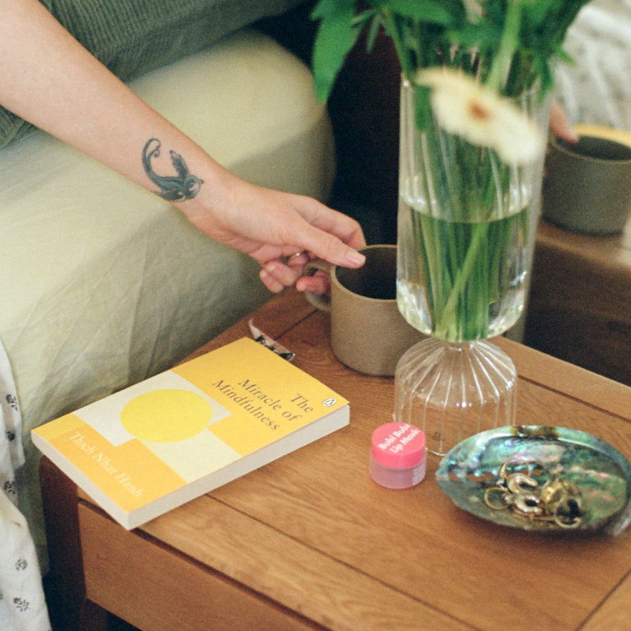A lifestyle scene showing a hand resting near a book titled 'The Miracle of Mindfulness' by Thich Nhat Hanh on a wooden bedside table. The book is yellow and white, next to a glass vase with green stems, a small pink jar, and a decorative dish.