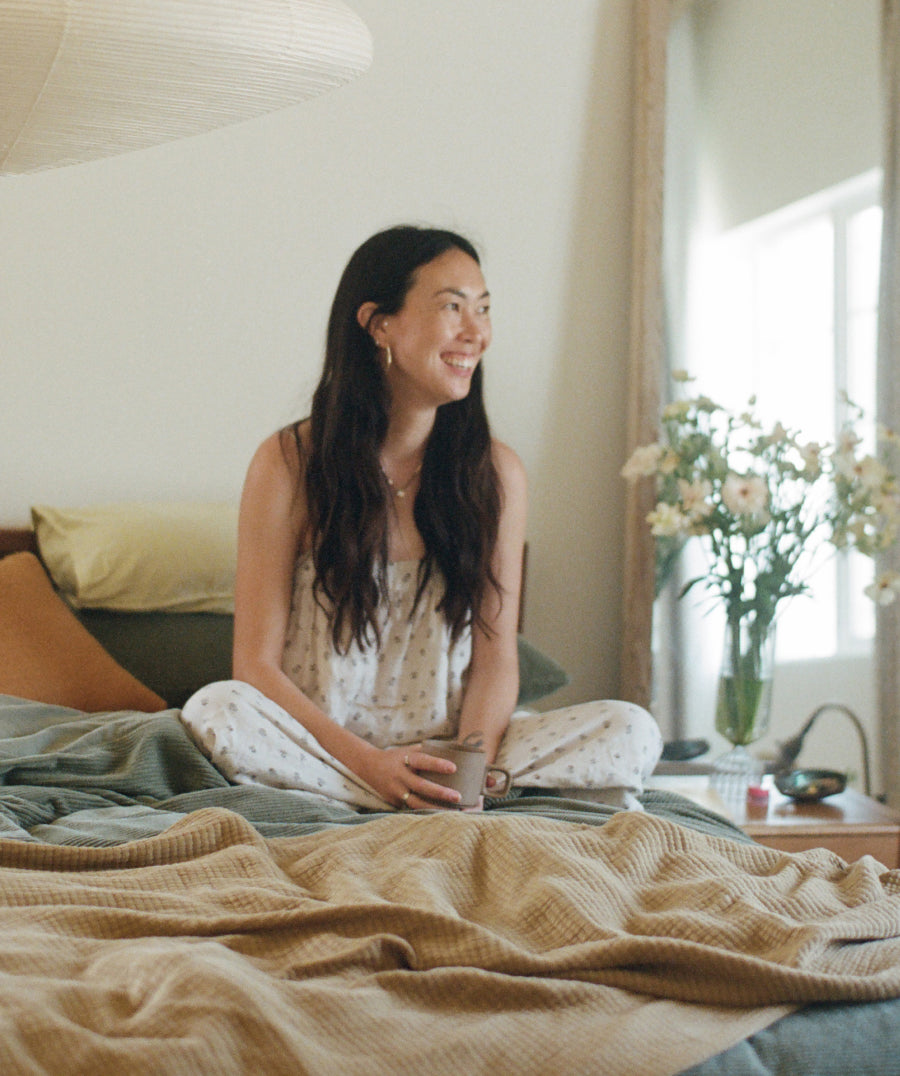 A woman sits smiling on a bed in a softly lit bedroom, wearing a floral sleep dress. A beige blanket covers the bed, with pillows, a lamp, and a vase of flowers visible in the room.