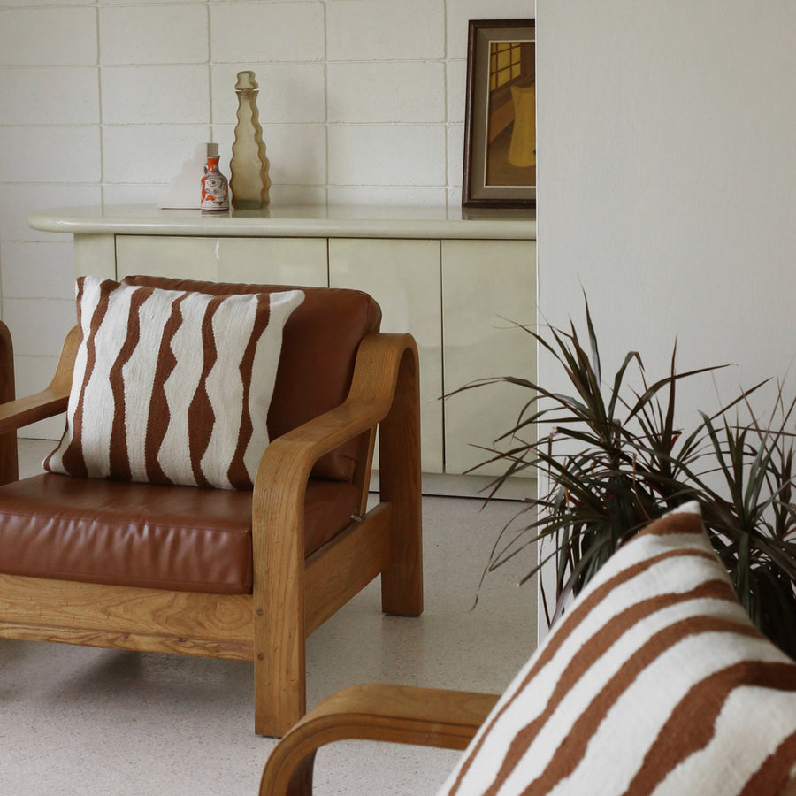 A modern living room setting with  a leather and wood chair featuring the Paso throw pillow with wavy thick stripes against a cream background